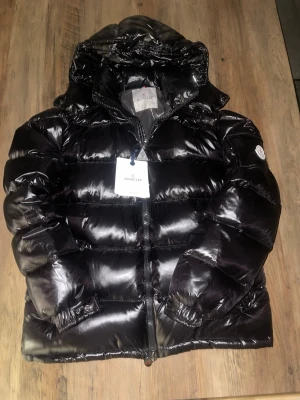 Moncler jacka - Svart Moncler maya med glansig finish och klassisk pufferdesign. Jackan har huva, dragkedja framtill och Moncler-logga på ärmen. Perfekt för kalla vinterdagar och riktigt varm tack vare dunfyllningen. Har en justerbar midja och är nyskick.