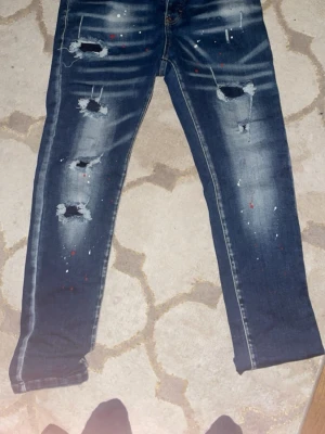 Mörkblå distressed jeans med färgstänk - Säljer ett par mörkblå jeans med slitna detaljer och stora hål på framsidan. Jeansen har vita och röda färgstänk samt blekta partier både fram och bak. Klassisk femficksmodell med raka ben och cool streetstil.