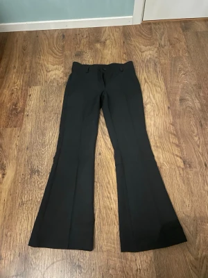 Svarta utsvängda kostymbyxor RYVLS - Svarta kostymbyxor från RYVLS med utsvängda ben och klassisk passform. Byxorna har hällor för skärp och pressveck framtill. Perfekta för en stilren look och lätta att matcha med olika toppar.