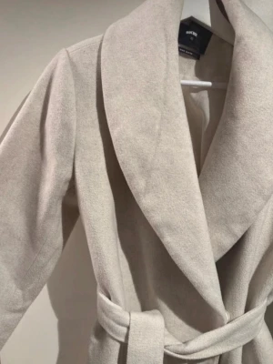 Ljusbeige kappa med skärp Bik Bok - Stilren ljusbeige kappa från Bik Bok med bred sjalkrage och knytskärp i midjan. Kappan har en rak passform och är tillverkad i ett mjukt tyg som ger en lyxig känsla. Perfekt för att lyfta din outfit under kyliga dagar.