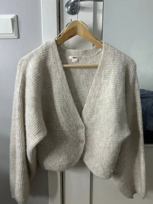 Beige stickad kofta med v-ringning - Mysig beige stickad kofta med v-ringning och två knappar framtill. Koftan har en loose passform och långa ärmar, perfekt för lager-på-lager-stil. Enkel och stilren design som passar till det mesta.