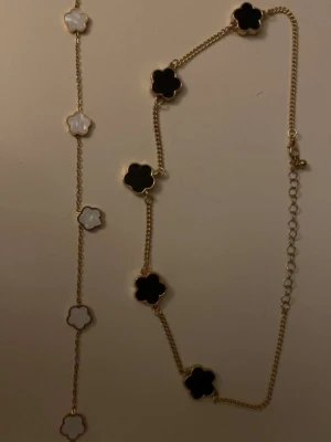 Guldhalsband med blommor i svart och vitt - Snyggt halsband med guldfärgad kedja och blommor i svart och vit pärlemor. Blommorna har en fyrklöverform och är jämnt placerade längs kedjan. Perfekt accessoar för att lyfta din outfit med en elegant touch.