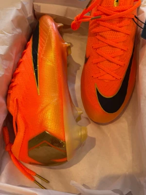 Nike Mercurial Vapor 16 orange fotbollsskor - Snygga Nike Mercurial Vapor 15 fotbollsskor i neonorange med svart swoosh och gulddetaljer. Ovandelen är i syntetmaterial med ventilerande mesh och sulan har en glansig, iriserande finish. Skorna har snörning och aggressivt dobbsystem för max grepp på planen.