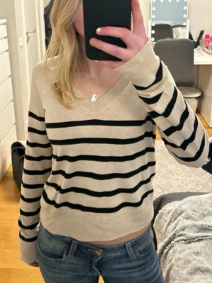 Beige randig tröja med V-ringning - Snygg beige tröja med svarta horisontella ränder och V-ringning. Långärmad och i mjukt material, perfekt för en chill och stilren look. Passar till jeans eller kjol för en avslappnad vibe.