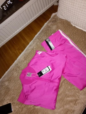 Rosa Adidas loungewear-set med prislapp  - Säljer ett fräscht rosa loungewear-set från Adidas med matchande t-shirt och byxor. Setet har vita detaljer på axlar och ben samt Adidas-logga på byxan. Perfekt för chill dagar hemma. Materialet känns mjukt och bekvämt.