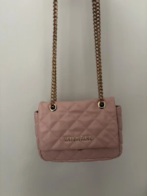 Rosa quiltad axelväska från Valentino - Snygg rosa axelväska från Valentino med quiltad design och guldfärgade kedjehandtag. Väskan har ett elegant och stilrent utseende med logotypen framtill. Perfekt för dig som vill ha en trendig och chic accessoar.