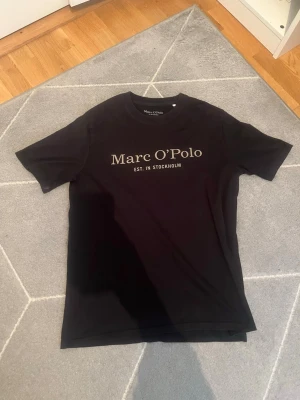 Svart Marc O'Polo t-shirt med tryck - Svart t-shirt från Marc O'Polo med rund hals och klassisk passform. Framsidan har ett stort guld tryck med texten 'Marc O'Polo EST. IN STOCKHOLM'. Perfekt för en stilren och enkel look.