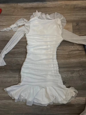 Vit volangklänning med långa ärmar - Supersöt vit kortklänning med långa ärmar i ett lätt, transparent material. Klänningen har volangdetaljer vid halsringningen och nederkanten samt rynkade partier längs hela kroppen för en snygg passform. Perfekt för dig som gillar romantisk och trendig stil.