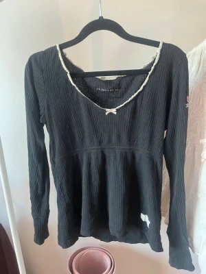 Svart ribbad topp med rosett - Svart långärmad ribbad topp med vit spetskant och liten rosett vid ringningen. Från Odd Molly.