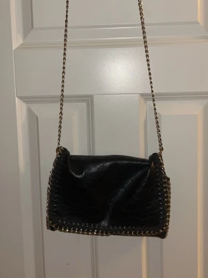 Svart axelväska med kedjerem - Snygg svart axelväska i fejkläder med kedjerem och dekorativa sömmar längs kanterna. Väskan har ett stort huvudfack och en innerficka med dragkedja. Perfekt för dig som gillar en stilren look. Passar perfekt till vardagens alla outfits. Finns dock ett hål som man ser på sista bilden, men inget som påverkar intrycket. 