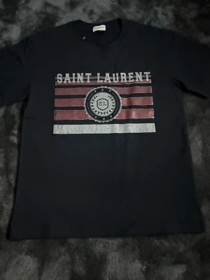 Svart Saint Laurent t-shirt - Cool svart t-shirt från Saint Laurent med tryck i vitt och rött framtill. T-shirten har klassisk passform och korta ärmar. Materialet känns mjukt och skönt, perfekt till vardags. Snygg logga och grafiskt mönster på bröstet.