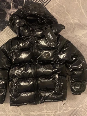 Svart pufferjacka från Trapstar - Säljer en glansig svart pufferjacka från Trapstar med stor huva och feta broderade Trapstar-bokstäver på ryggen. Jackan har två stora fickor framtill, dragkedja och ett runt Trapstar-märke på bröstet. Perfekt för kalla vinterdagar och riktigt street.