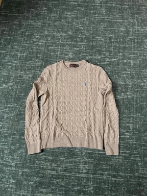 Beige kabelstickad tröja Morris - Kabelstickad beige tröja från Morris  med rund halsringning och lång ärm. Tröjan har ett litet blått broderat logomärke på bröstet och ribbstickade muddar vid ärmslut och nederkant. Perfekt för en stilren och klassisk look.