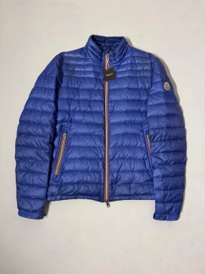 Moncler Daniel Down Jacket - Moncler Daniel jacka en i sällsynt blå färg. Riktigt fint skick, endast väldigt små vattenfläckar på vissa ställen som endast syns om man tittar noga samt en liten reva/hål lagat snyggt med laglapp. Storlek 3 dvs som en medium.