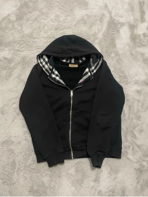 Svart hoodie med dragkedja Burberry - Svart hoodie från Burberry med dragkedja och klassiskt rutigt foder i huvan och insidan. Hoodien har två fickor framtill och ribbade muddar vid ärmslut och nederkant. Perfekt för en stilren och trendig look. KOM INTE MED DUMMA PRIS FÖRSLAG