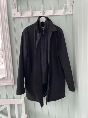 NN07 Svart Vinterrock Herr Strl XL – Avtagbar Innerjacka - Svart vinterrock från NN07 i storlek XL med rak, stilren passform och knäppning framtill. Rocken har en avtagbar quiltad innerjacka med hög krage och dragkedja som ger extra värme och flexibilitet. Diskreta sidofickor. En funktionell och elegant modell som passar både till vardag och mer uppklädda tillfällen. Mycket fint skick.