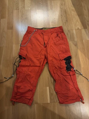 Orange cargobyxor med kedja och snörning - Säljer ett par orange cargobyxor med cool kedjedetalj i midjan, svarta snörningar på sidorna och flera fickor. Byxorna har dragkedjor vid bensluten och är perfekta för en streetwear-look. Materialet är bomull och de har en avslappnad passform.