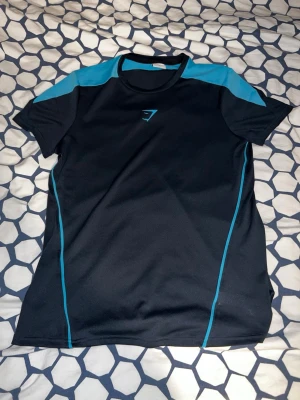 Svart och blå träningströja Gymshark - Snygg svart träningströja från Gymshark med blå detaljer på axlar och sidor. Tillverkad i ett lätt och snabbtorkande material som passar perfekt för gymmet eller löpning. Tröjan har rund hals och korta ärmar samt Gymshark-logga på bröstet.