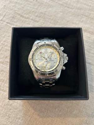 Silver Chrono Diver Herrklocka - Breitling Chronograph med boett och armband i stål. Urtavlan är vit med tre underrattar och datumvisning. Klockan har en vridbar bezel och fungerande kronografknappar på sidan av boetten. Baksidan är märkt "STAINLESS STEEL BACK 3ATM WATER. Trippel A Replik@. Saknar bara batteri. 