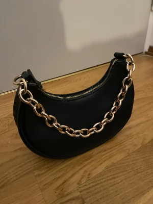 Svart axelväska med guldkedja - Snygg svart handväska i halvmåneformad modell med bred guldfärgad kedja som axelband. Väskan har dragkedja upptill och är gjord i slätt konstläder. Perfekt accessoar för att lyfta din outfit.