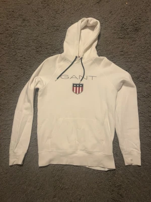 Vit hoodie från GANT med logga - Snygg vit hoodie från GANT med stor huva och dragsnören. På bröstet finns GANT-loggan och ett broderat sköldmärke i rött, vitt och blått. Hoodien har en klassisk känguruficka framtill och ribbade muddar vid ärmar och nederkant. Perfekt för en clean och sportig look.
