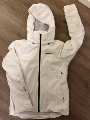 Vit vindjacka från Peak Performance - Snygg vit jacka från Peak Performance med svart dragkedja och logga på bröstet. Jackan har huva, justerbara detaljer och två sidofickor med dragkedja. Tillverkad i ett slitstarkt, vattenavvisande material som passar perfekt