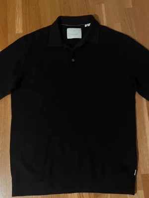 Svart pikétröja från Jack & Jones - Klassisk svart pikétröja från Jack & Jones med krage och knappslå. Perfekt till jeans eller chinos för en clean look. Tröjan har korta ärmar och är tillverkad i mjukt material.