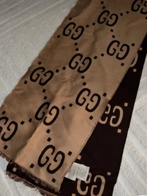 Beige-brun halsduk från Gucci - Snygg halsduk från Gucci i beige och mörkbrun med det klassiska GG-mönstret över hela tyget. Halsduken har fransade kanter och är tillverkad i ett mjukt material som känns lyxigt. Perfekt accessoar för att lyfta din outfit.