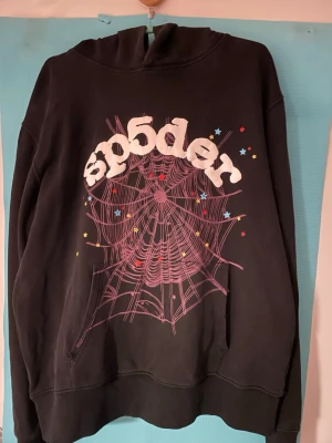 Svart hoodie från Sp5der med tryck - Svart hoodie från Sp5der med stort vitt logotryck och rosa spindelnätsgrafik på framsidan. Färgglada små stjärnor ger extra detaljer. OBS Litet hål i armhålan som inte syns när man har på sig den.