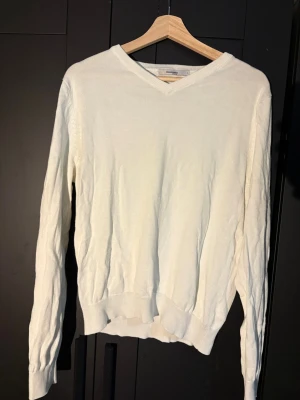 Vit stickad tröja med V-ringning - Klassisk vit stickad tröja med V-ringad hals och långa ärmar. Tröjan har ribbade muddar vid ärmslut och nederkant. Perfekt att matcha med jeans eller kjol för en clean och enkel look.
