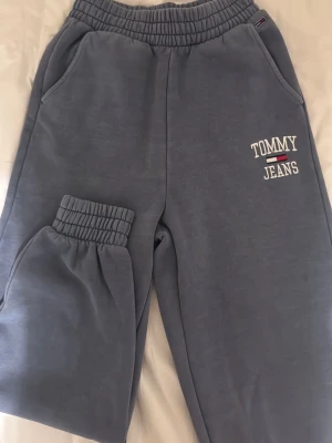 Blå/grå mjukisbyxor Tommy Jeans XS - Blå/grå mjukisbyxor från Tommy Jeans med broderad logga på benet. Byxorna har resår i midjan och vid bensluten, samt två sidofickor. Insidan är fodrad med mjuk fleece för extra komfort. Perfekta för chill och avslappnade dagar.