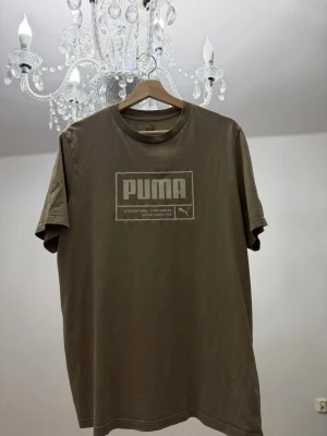 Brun/beighe t-shirt från Puma - Snygg brun/beighe  t-shirt från Puma med klassisk logga framtill. T-shirten har rund halsringning och korta ärmar, perfekt för en avslappnad och sportig stil. Tillverkad i mjuk bomull som känns skön mot huden.