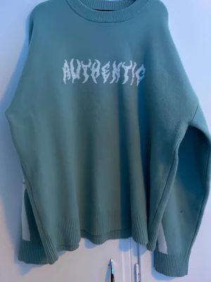 Ljusblå stickad tröja med skelettmönster - Cool stickad tröja i ljusblå med vit text 'AUTHENTIC' på bröstet och stort skelettmönster på ryggen. Oversized passform, ribbade muddar och rund hals. Perfekt för dig som gillar streetwear och unika detaljer.
