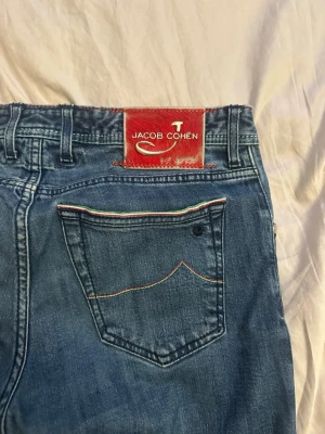 Blå jeans från Jacob Cohen - Snygga blå jeans från Jacob Cohen med klassisk femficksmodell och röd patch bak. Jeansen har raka ben och detaljerade sömmar, samt en liten färgdetalj på bakfickan. Tillverkade i mjukt denimtyg som ger en skön känsla. Pyttet slitet skav i skrevet som man inte märker av. Nypris över 5000
