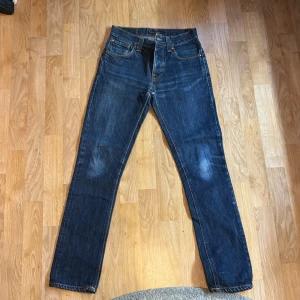 Mörkblå slim jeans från Nudie - Säljer ett par mörkblå jeans från Nudie med klassisk rak passform och fem fickor. Jeansen har kontrastsömmar och subtila slitningar på framsidan. Tillverkade i kraftig denim med Nudie-logga på bakfickan. Perfekta för en avslappnad och stilren look.
