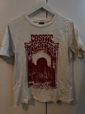 Cosmic Dreamers Soul Searcher T-shirt - Snygg vit t-shirt från Cosmic Dreamers med ett stort rödbrunt tryck framtill där det står 'Soul Searcher'. T-shirten har klassisk passform, rund hals och korta ärmar. Perfekt för dig som gillar retroinspirerad stil och coola prints.