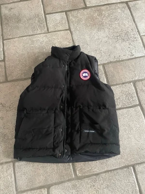 Svart dunväst från Canada Goose - Säljer en svart dunväst från Canada Goose med hög krage och två stora fickor framtill. Västen har dragkedja och tryckknappar samt den klassiska Canada Goose-loggan på bröstet. Perfekt för lager-på-lager och riktigt varm under kalla dagar.obs städas och tvättas inan jag skickar 