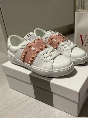 Valentino Garavani sneakers med nitar - Snygga vita sneakers från Valentino Garavani med breda ljusrosa band. Använda typ en gång i bra skick storleken passa inte mig då jag har 36 och dessa är 37.