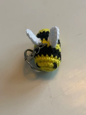 Virkad bi nyckelring - Söt handvirkad nyckelring formad som ett bi i gult och svart med vita vingar. Perfekt att hänga på väskan eller nyckelknippan för en personlig touch. 