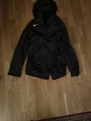 Svart Nike vindjacka med huva - Snygg svart vindjacka från Nike med huva och dragkedja framtill. Jackan har en vit Nike-logga på bröstet och är tillverkad i ett lätt material som skyddar mot vind. Perfekt för dig som vill ha en sportig och stilren look.