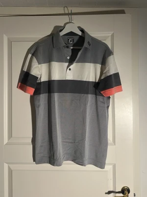 Randig pikétröja från FootJoy - Snygg pikétröja från FootJoy med breda ränder i grått, vitt, svart och en touch av orange på ärmarna. Klassisk krage med knappar framtill och korta ärmar. Tillverkad i ett mjukt material som känns skönt mot huden.