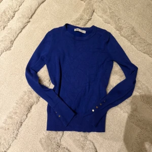 Kornblå stickad tröja från Zara - Snygg kornblå stickad tröja från Zara med rund halsringning och långa ärmar. Tröjan har dekorativa guldfärgade knappar vid ärmsluten och är tillverkad i ett mjukt, tunt material som sitter skönt på kroppen.