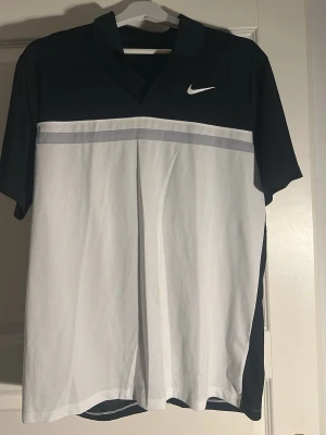 Nike svartvit pikétröja - Sportig pikétröja från Nike i svart och vitt med grå detalj över bröstet. Tröjan har korta ärmar, klassisk krage och en vit Nike-logga på bröstet. Perfekt för träning eller golf. Tillverkad i ett lätt och ventilerande material.