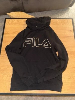 Svart hoodie från FILA med logga - Svart hoodie från FILA med stor vit logga på bröstet. Klassisk modell med huva och magficka, ribbade muddar vid ärmslut och nederkant. Tillverkad i mjukt material som känns skönt mot huden. Perfekt för en avslappnad och sportig stil.
