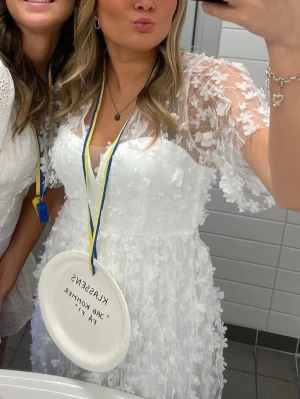 Vit blommig kortklänning - Superfin vit kortklänning med blommiga 3D-detaljer. Klänningen har v-ringning, genomskinliga ärmar och volangkant. Perfekt för student eller sommarens alla tillfällen. Använd en gång (försiktigt använd), inga tecken på användning!