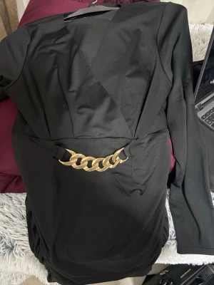 Svart omlottklänning med guldkedja - Snygg svart omlottklänning med lång ärm och en stor guldfärgad kedjedetalj i midjan. Klänningen har en elegant v-ringning och markerad midja som ger en stilren siluett. Perfekt för dig som vill sticka ut med en cool detalj.