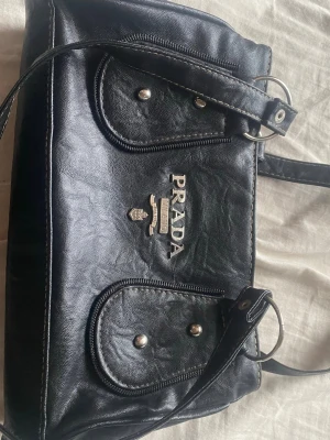 Svart liknande handväska - Snygg svart handväska, inget märke! Två framfickor med lock och silverfärgade detaljer. Väskan har justerbar axelrem och tydlig logga i metall framtill. Perfekt för dig som vill ha en klassisk och stilren accessoar.