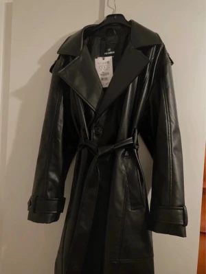 Svart trenchcoat i skinnimitation Pull&Bear - Ny, stilren svart trenchcoat i fuskläder från Pull&Bear. Jackan har bred krage, knytskärp i midjan och detaljerade ärmslut med spännen. Perfekt för dig som vill ha en edgy och trendig look. Långa ärmar och klassisk passform. Står storlek S men är overzised. Skriv för eventuella frågor. 
