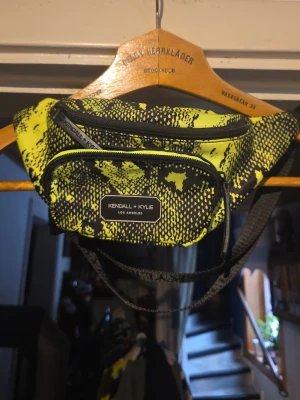 Neongul magväska Kendall + Kylie - Snygg magväska från Kendall + Kylie i neongult och svart med coolt mönster. Väskan har två dragkedjefack och justerbart band. Perfekt för att bära det viktigaste och ger en riktigt trendig streetstyle-look.