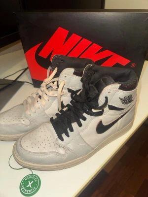 Nike Air Jordan 1 High NYC TO PARIS - Snygga Nike Air Jordan 1 High OG sneakers i grått och svart läder med klassisk svart swoosh och Air Jordan-logga på sidan. Skorna har en hög siluett och olika färg på skosnörena, ett par vita och ett par svarta. Perfekta för dig som gillar streetwear och ikonisk design.strk 44. Passar mig som vanligt har 43